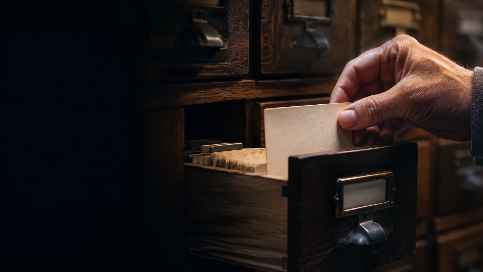 A hand placing a blank archival card into a wooden library card catalog drawer.